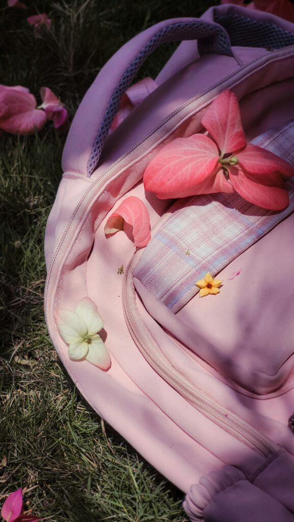 A pink backpack adorned with vibrant petals, set on green grass under sunlight, creating a serene outdoor scene.