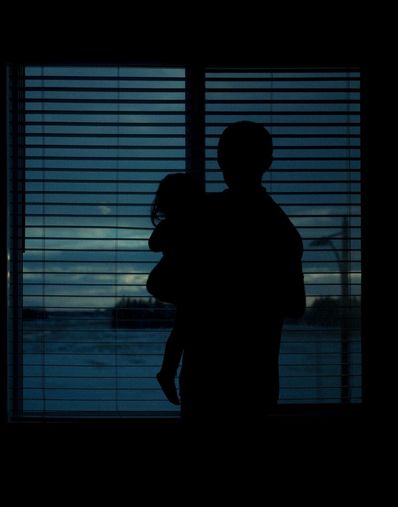 A dramatic silhouette of a parent holding a child against a window with blinds, creating a serene indoor scene.